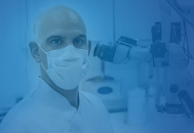 A male dentist wearing scrubs and a face mask, looking into the camera