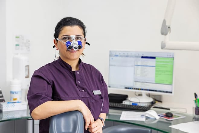 A dentist in purple scrubs leans on the chair wearing loupes looking at the camera