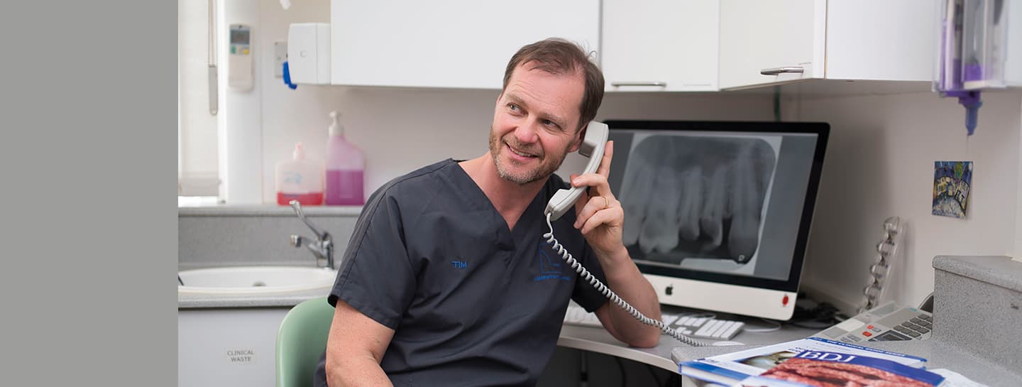 A dentist wearing dark grey scrubs is on the phone at a desk in a dental surgery