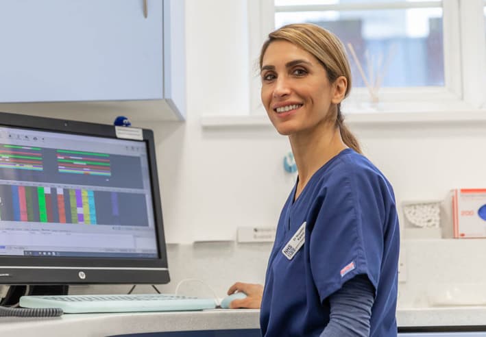 A female dentist in blue scrubs sitting in front of a computer, smiling at the camera 