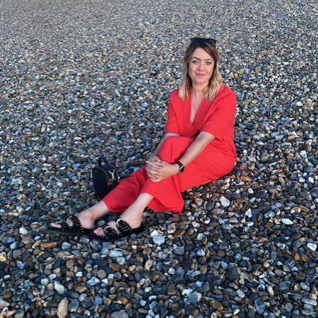 Lauren Firth wearing a red dress sitting on a pebble beach